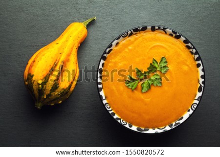 Roasted butternut squash or pumpkin soup, bowl of soup decorated with fresh parsley leaves, next to an orange pear shaped ornamental gourd on black slate rock board surface, flat lay overhead top view
