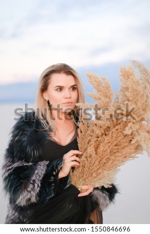 Portrait of girl wearing fur coat with dried plants bouquet in snowed field background. Concept of winter photo session and beauty, fashionable female person.
