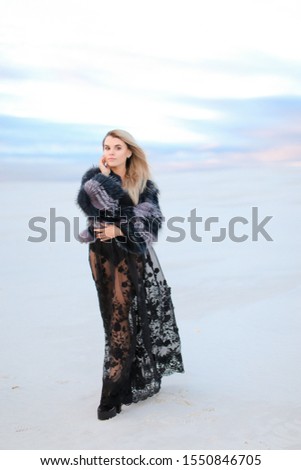 Young female person wearing black dress standing in white snowed steppe background. Concept of fashion and winter photo session.