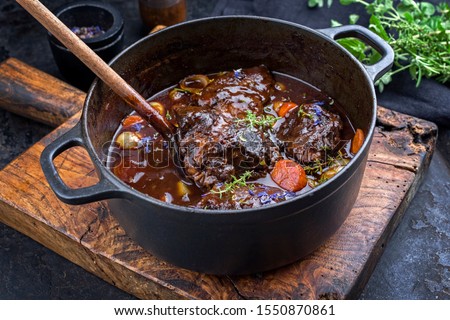 Traditional German braised beef cheeks in brown red wine sauce with carrots and onions offered as closeup in a cast iron Dutch oven on an old rustic cutting board  Royalty-Free Stock Photo #1550870861