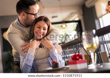 Couple on a date in restaurant, celebrating woman's birthday; man standing behind a woman and hugging her after giving her a birthday present and a little surprise birthday cake