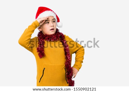 Little boy celebrating christmas day wearing a santa hat isolated looking far away keeping hand on forehead.