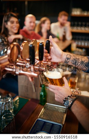 Cropped picture of tattooed barman pouring beer in glass. In background is group of friends sitting at counter. Pub interior.