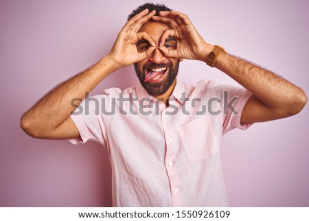 Young indian man wearing casual shirt standing over isolated pink background doing ok gesture like binoculars sticking tongue out, eyes looking through fingers. Crazy expression.