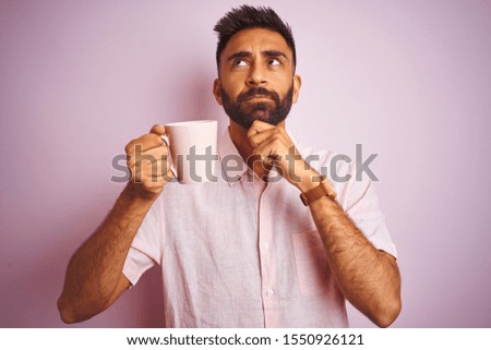 Young indian man wearing shirt drinking cup of coffee standing over isolated pink background serious face thinking about question, very confused idea