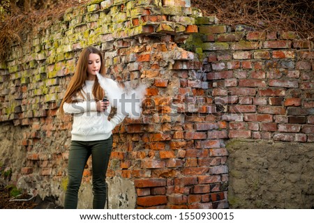 Vaping teenager. Young pretty white caucasian teenage girl with problem skin smoking an electronic cigarette the ruined brick wall on the street in the autumn. Bad habit. Vape activity.