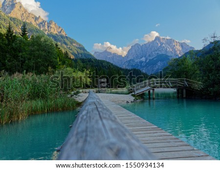 Photo of Lake jasna with wooden fence of the bridge across the first lake leading into the photo. In the background there are mt Prisojnik on the right and mt Škrlatica to the left. 