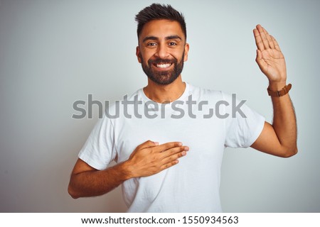 Young indian man wearing t-shirt standing over isolated white background smiling swearing with hand on chest and fingers up, making a loyalty promise oath Royalty-Free Stock Photo #1550934563