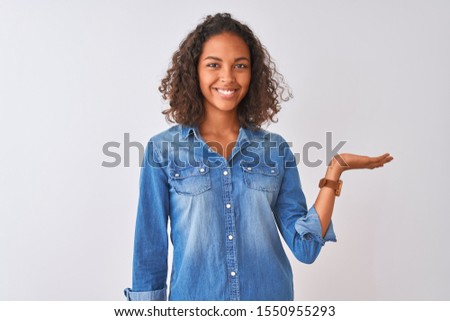 Young brazilian woman wearing denim shirt standing over isolated white background smiling cheerful presenting and pointing with palm of hand looking at the camera.