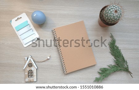 A top view mockup with a blue candle, a little house Christmas decoration, a cactus flower, a branch of evergreen tree and a blank notebook on the wooden background, flat lay