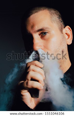 Stock vertical photography. A young man with blue eyes and a shaved head throws a large cloud of steam from his electronic cigarette through his nose.
