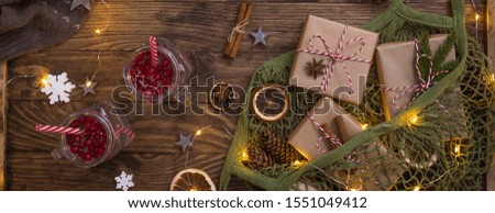 New Year Christmas composition with hot cranberry tea in glass mugs, net mesh bag with wrapped presents, cones, cinnamon, dried oranges, woolen sweater and lights on wooden background, banner