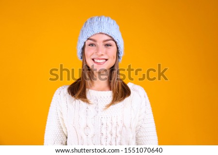 Close up portrait of a young beautiful woman with light make up on, wearing knitted sweater & blue woolen beanie. Attractive female in winter knitwear outfit, isolated on yellow background. Copy space
