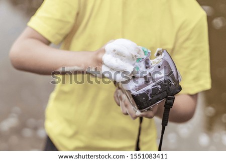 A boy in a yellow t-shirt and shorts is standing near the water in nature and washing a camera with soap and foam. The concept child is a bully. photographers day, joke