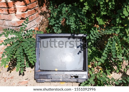 An old broken black television lays in garbage and bushes. Sun light reflection.