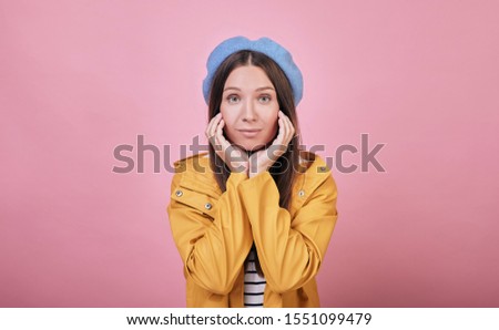 Cool girl in a gentle blue beret, a striped blouse and a yellow rain jacket smiles cute against a pink background. Arms apart and she smiling.