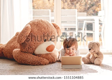 Cute little child watching cartoons on digital tablet device lying on floor with two soft teddy bear toys at home. Modern childhood. Quarantine and isolation period concept. Child alone at home