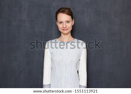 Studio half-length portrait of satisfied friendly young woman wearing light-colored jumper, looking kindly at camera, smiling, being in good mood, happy and cheerful, standing against gray background