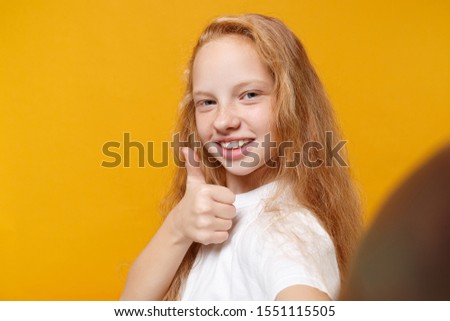 Close up of little ginger kid girl 12-13 years old in t-shirt isolated on bright yellow background. Childhood lifestyle concept. Mock up copy space. Doing selfie shot on mobile phone showing thumb up