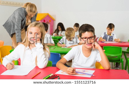 Portrait of schoolgirl and schoolboy at desk with classmates and teacher on background