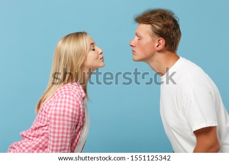 Young pretty couple two friends guy girl in white pink empty blank design t-shirts posing isolated on pastel blue background. People lifestyle concept. Mock up copy space. Kissing keeping eyes closed