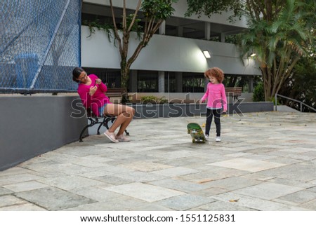 Caucasian girl playing skateboard and smiling happily against and mother taking pictures with cellphone