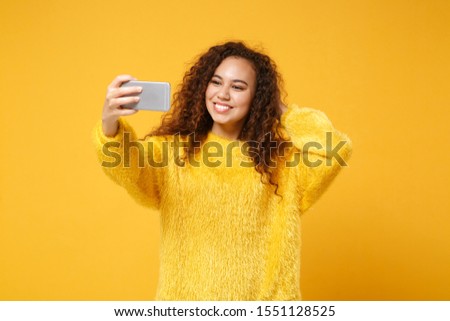 Smiling young african american girl in fur sweater posing isolated on yellow orange wall background, studio portrait. People lifestyle concept. Mock up copy space. Doing selfie shot on mobile phone