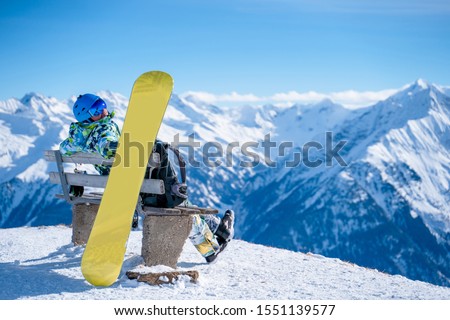 Image from back of athlete man sitting on bench next to snowboard in ski resort.