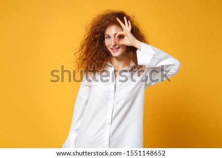 Young redhead woman girl in casual white shirt posing isolated on yellow orange wall background. People lifestyle concept. Mock up copy space. Holding hand near eye imitating binoculars or eyeglasses