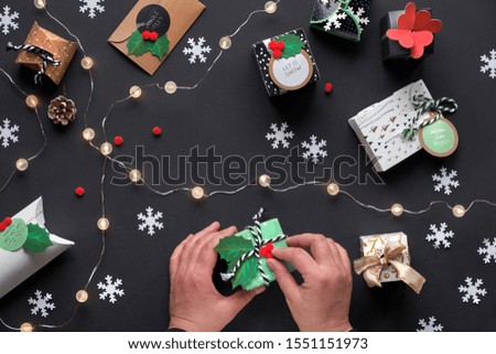 New Year or Christmas presents wrapped in various paper gift boxes with tags. Hands decorating box with holly. Festive flat lay, top view with light garland, green alarm and snowflakes on black paper.
