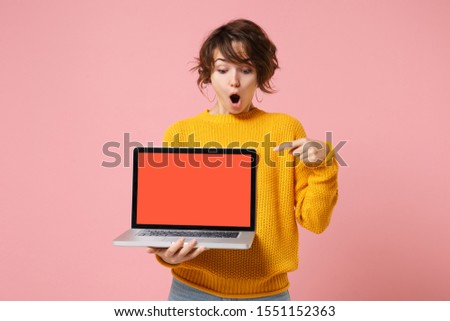 Shocked young brunette woman girl in yellow sweater posing isolated on pink background. People lifestyle concept. Mock up copy space. Point index finger on laptop pc computer with blank empty screen
