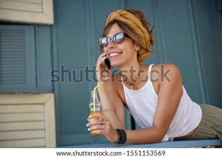 Outdoor photo of young brunette woman in vintage sunglasses wearing casual clothes and mustard headband, posing over summer terrace and drinking lemonade, looking ahead and smiling happily