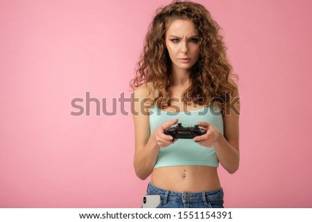 Pretty gamer girl with curly hair holding gaming controller and express positive emotion on pink background