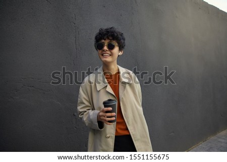 Plasant looking attractive curly brunette woman with casual hairstyle holding takeaway coffee and smiling slightly, dressed in trendy clothes and sunglasses, posing over city environment