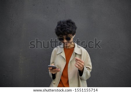 Cheerful pretty young curly brunette woman with short haircut keeping mobile phone in hand while posing over black city wall, wearing stylish clothes and sunglasses
