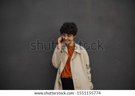 Outdoor shot of young curly brunette woman with short haircut in trendy outfit making call with her mobile phone while walking over city streets, posing over black urban wall in stylish clothes