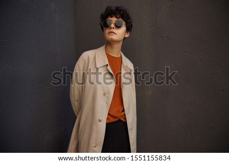 Fashionable young dark haired pretty female with short haircut standing over black urban wall an keeping hands behind her back, wearing trendy outfit and sunglasses