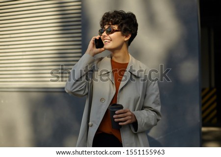 Pleasant looking cheerful young curly woman with casual hairstyle walking outside on sunny warm day, having pleasant talk on phone, wearing trendy clothes and stylish sunglasses
