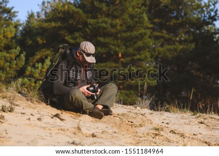 Mature man look at his photocamera while sitting at sand meadow inside park location. Man photographer looks at camera and checks shot on monitor.