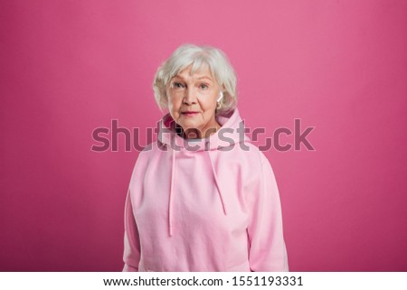 Picture of calm, peaceful and beautiful old lady look straight forward and pose on camera. Stand straight and wear hoody. Model with grey hair and wrinkles on face. Isolated over pink background