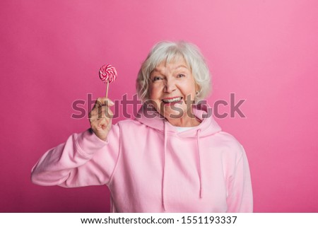 Cheerful attractive senior female model on picture. Hold lollipop in hand and smile. Beautiful old woman in stylish modern hoody. Isolated over pink background