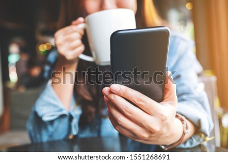 Closeup image of a woman holding  and using mobile phone while drinking coffee