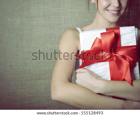 Portrait of attractive cheerful girl in sleeveless sports white shirt holding gift box with red bow over canvas background, toned and noise added