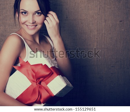Portrait of attractive cheerful girl in sleeveless sports white shirt holding gift box with red bow over canvas background, toned