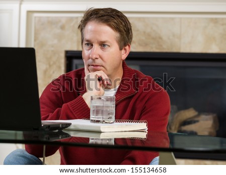 Photo of mature man, dressed casually while sitting down at glass table, working from home, looking at computer screen with fireplace in background 