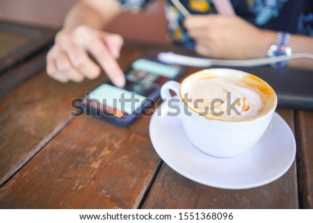Close up image of a cup of coffee on brown wooden table with woman sitting and using smartphone on the table, space for design and text