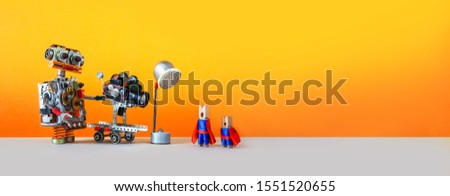 Comical art. Toy robot cameraman makes a movie about super heroes. Funny clothespin actors, robotic operator, camcorder spotlight. Filmmaking behind the scenes concept. Yellow background, empty space
