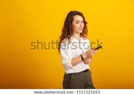 Beautiful young girl with a smartphone in hand looks at the camera and smiles standing isolated on yellow background