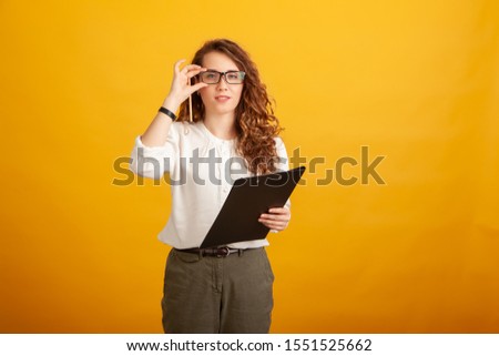 Beautiful young girl with a clipboard and a pencil touches her glasses and looks at the camera standing isolated on yellow background