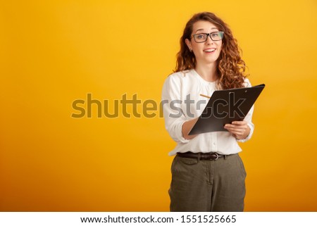Beautiful young girl in glasses with a clipboard smiles and looks in camera standing isolated on yellow background
